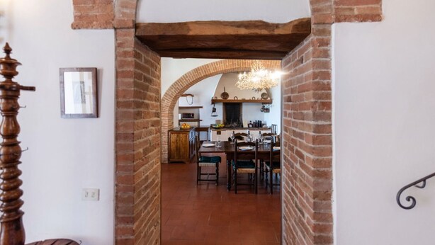 Un intérieur rustique avec une arche en briques donnant sur un espace repas agrémenté de meubles en bois et d’un lustre.