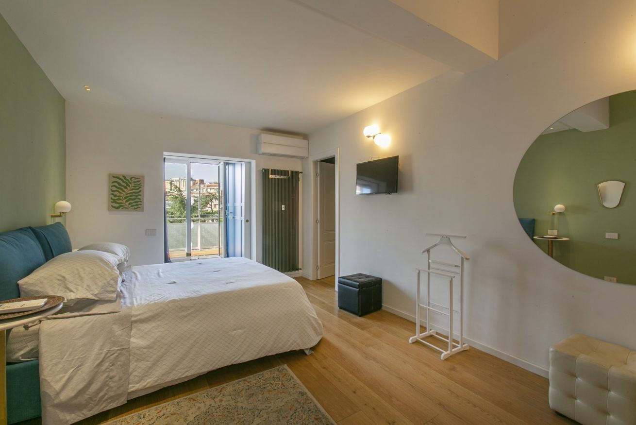 A bedroom with a bed, a small table, a chair, a hanging light fixture, and a view of the outside.