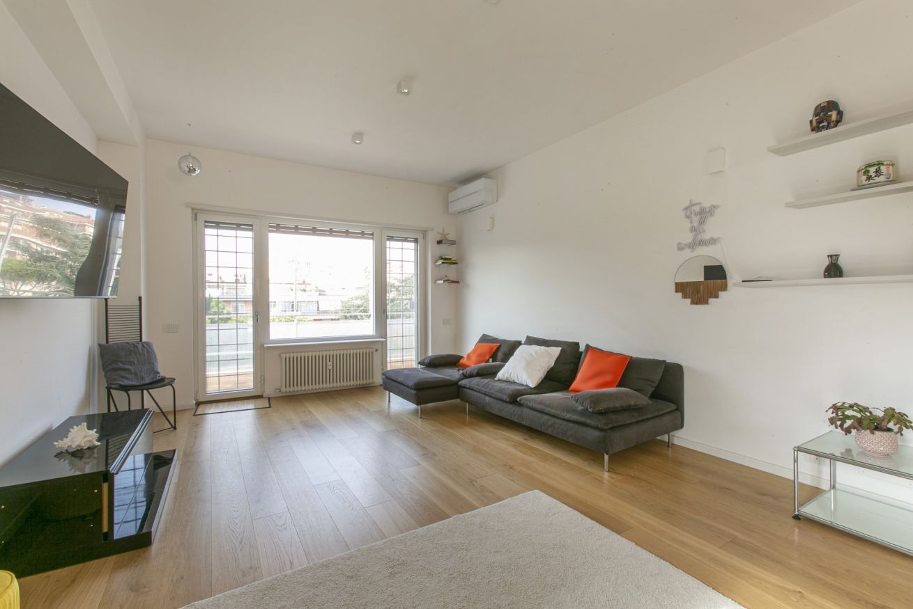 A spacious living room with a grey sectional sofa, a glass coffee table, and a large window with blinds.