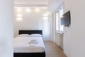 Een slaapkamer met een bed, witte muren, een wandtelevisie en een raam.