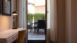 Habitación con escritorio, silla, lámpara y vistas a un patio exterior con sillas y seto.