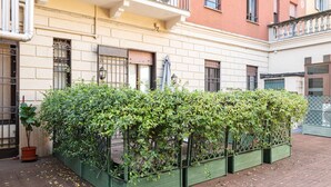 Un patio con una fila de macetas verdes que contienen arbustos frondosos, una planta en maceta y un suelo de baldosas.