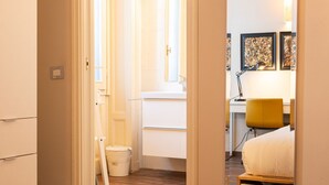 Una habitación de hotel compacta con una cama, un escritorio y una silla. Cuenta con un ventanal con cortinas, un lavamanos blanco y un cesto de basura.