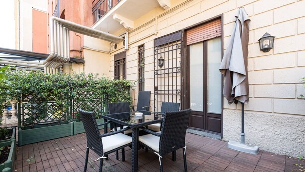 Un patio al aire libre con una mesa y sillas, cerca hay una valla verde y un edificio con balcón.
