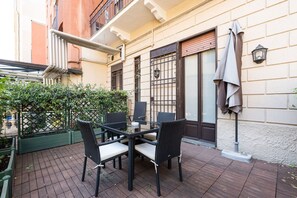 Une terrasse extérieure avec une table et des chaises, une clôture verte et un bâtiment doté d’un balcon.