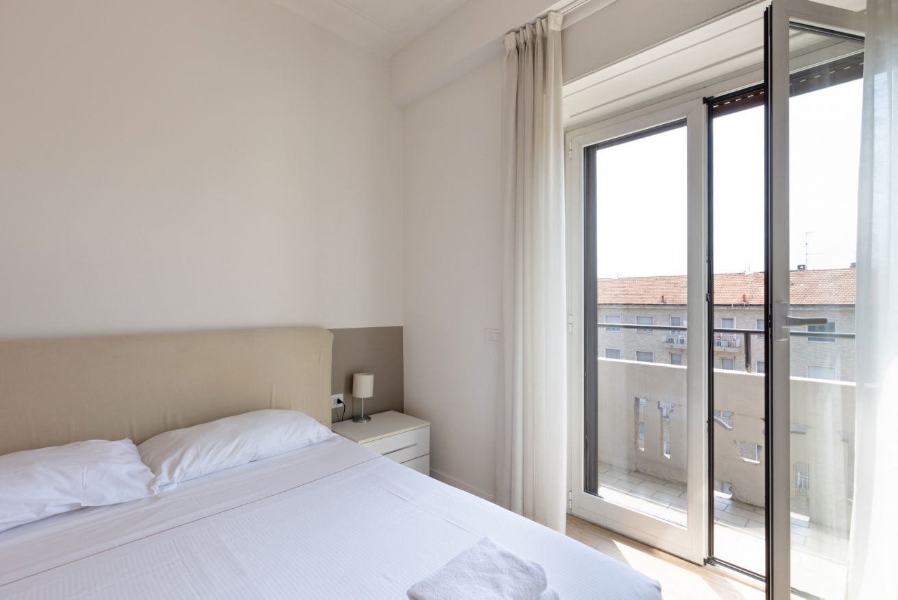 A hotel room with a bed, a nightstand, a lamp, and a sliding glass door to a balcony.