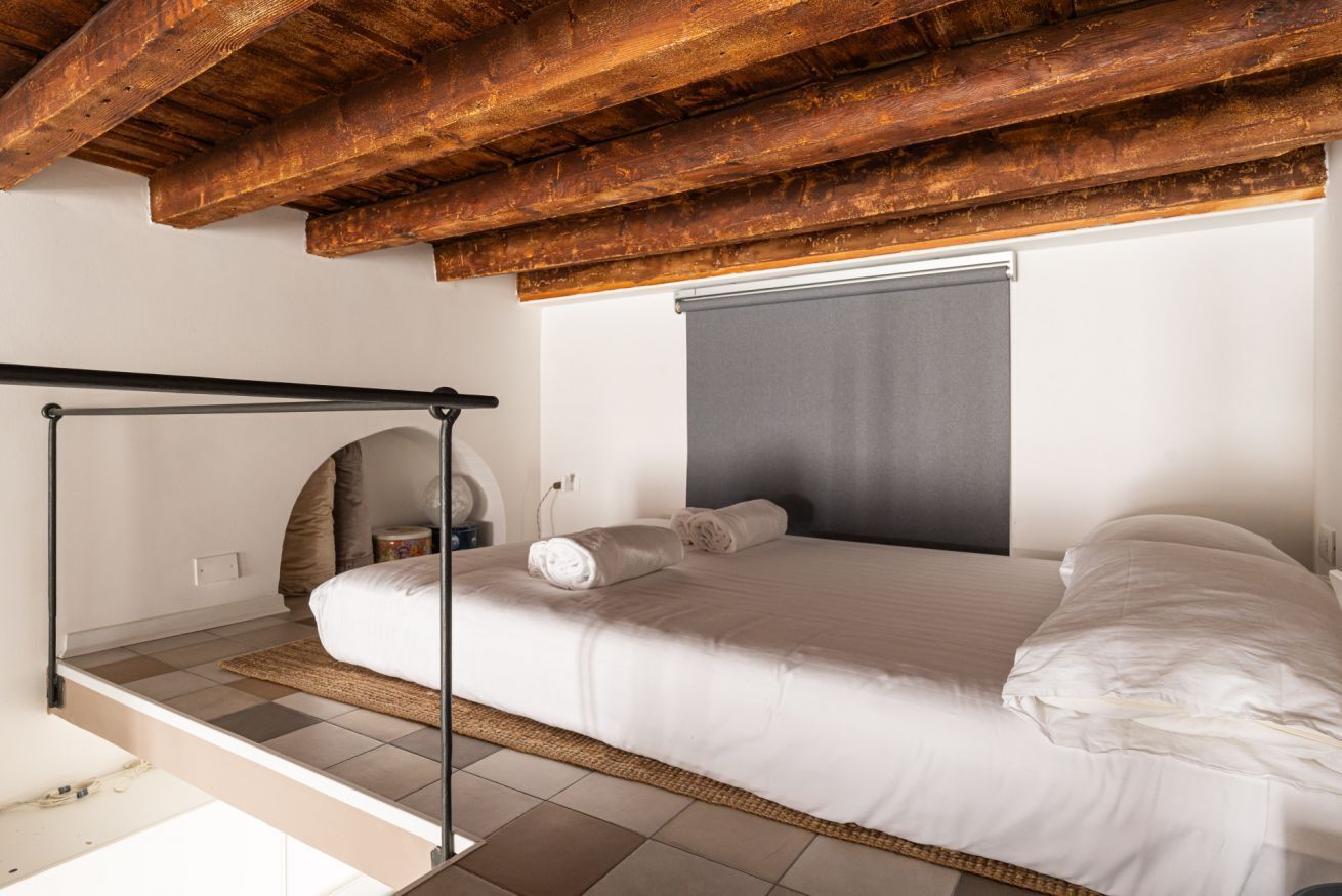 A bedroom with a bed, wooden ceiling beams, a window with a blind, and a small bedside table.