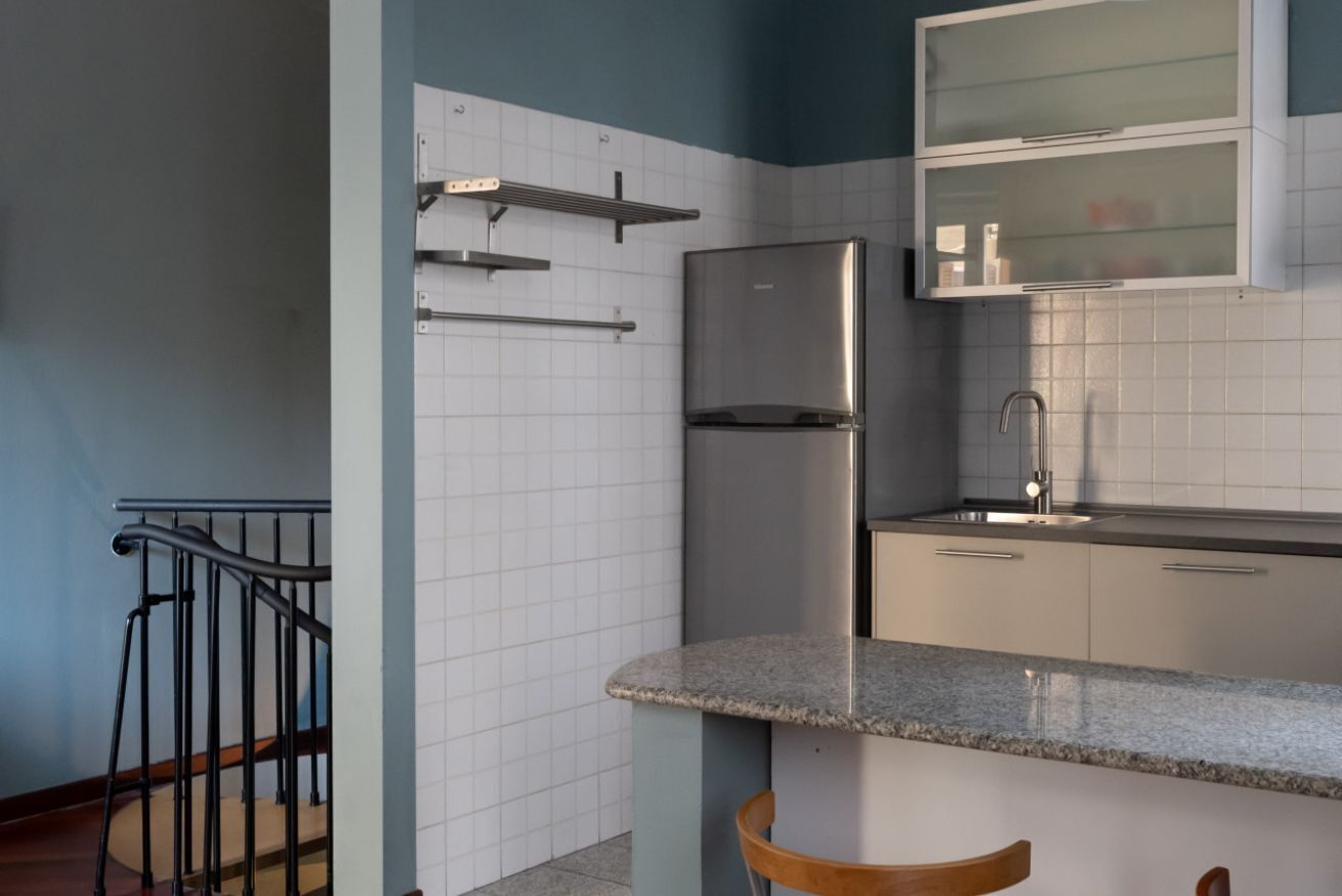 Une cuisine avec des murs recouverts de carreaux blancs, un réfrigérateur en acier inoxydable, un plan de travail en granit et une chaise en bois.