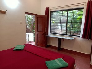 A room with a bed, a window with a view of greenery, a door, and a small table. - Namaste Yoga Farm (Kumta)