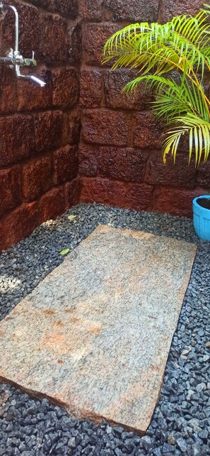 A stone-tiled shower area with a brick wall, a potted plant, and a blue container. - Namaste Yoga Farm (Kumta)