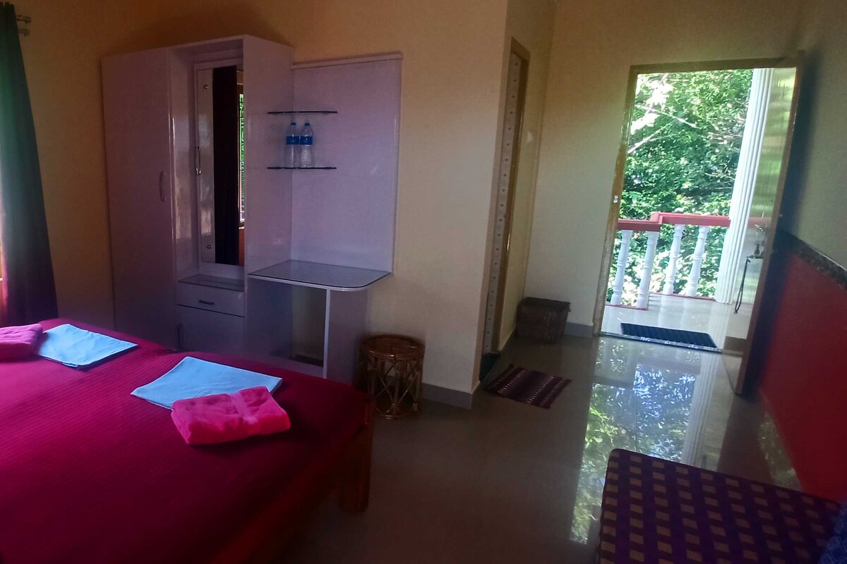 A room with a bed, a wardrobe, a small table, and a window with a view of greenery.