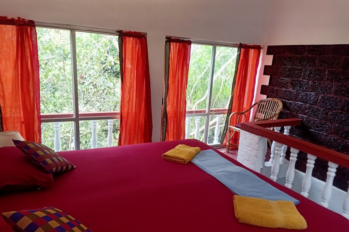 A bedroom with a large bed, two windows with red curtains, a white railing, and a brick wall.