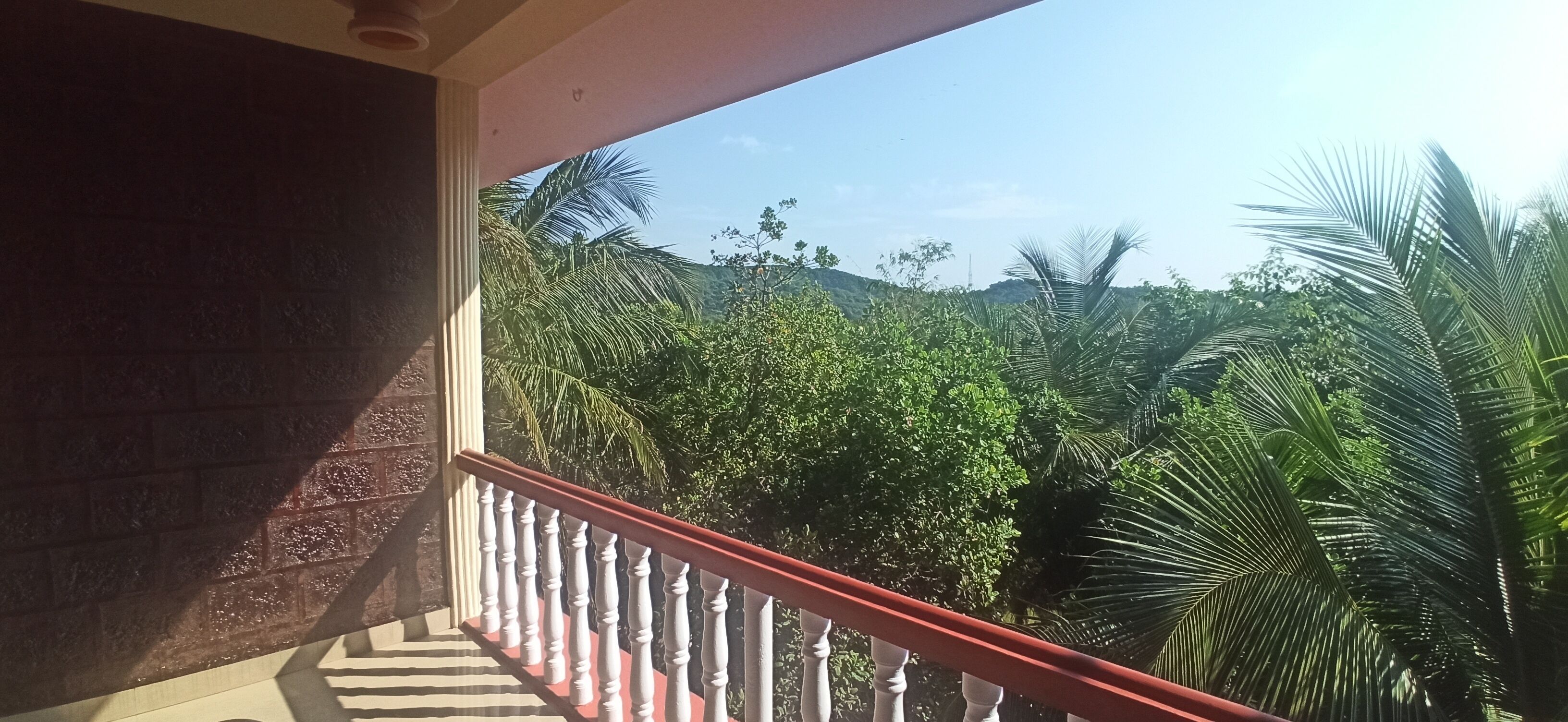 Un balcón con barandilla roja que da a un paisaje verde exuberante, con palmeras y colinas al fondo.