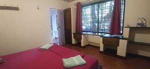 A room with two beds, a desk, a window with curtains, and a door leading to an outdoor area. - Namaste Yoga Farm (Kumta)
