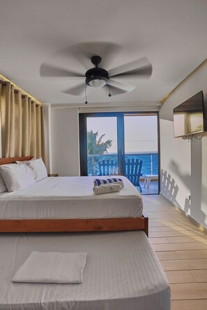 A hotel room with a bed, a ceiling fan, a TV mounted on the wall, and a view of the ocean through a sliding door.