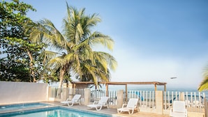 A pool area with lounge chairs, a pergola, and a view of the ocean.