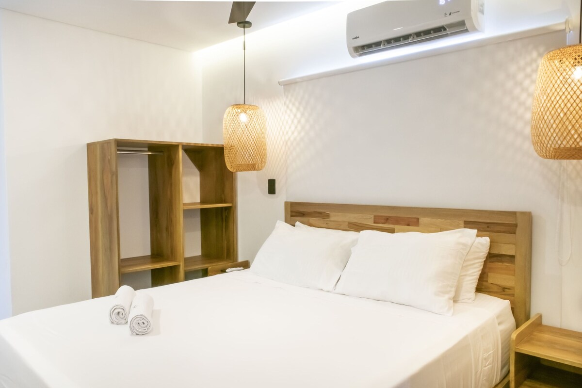 Un lit bien fait, avec du linge de lit blanc et des oreillers, une tête de lit en bois et une armoire en bois dotée d’étagères ouvertes.