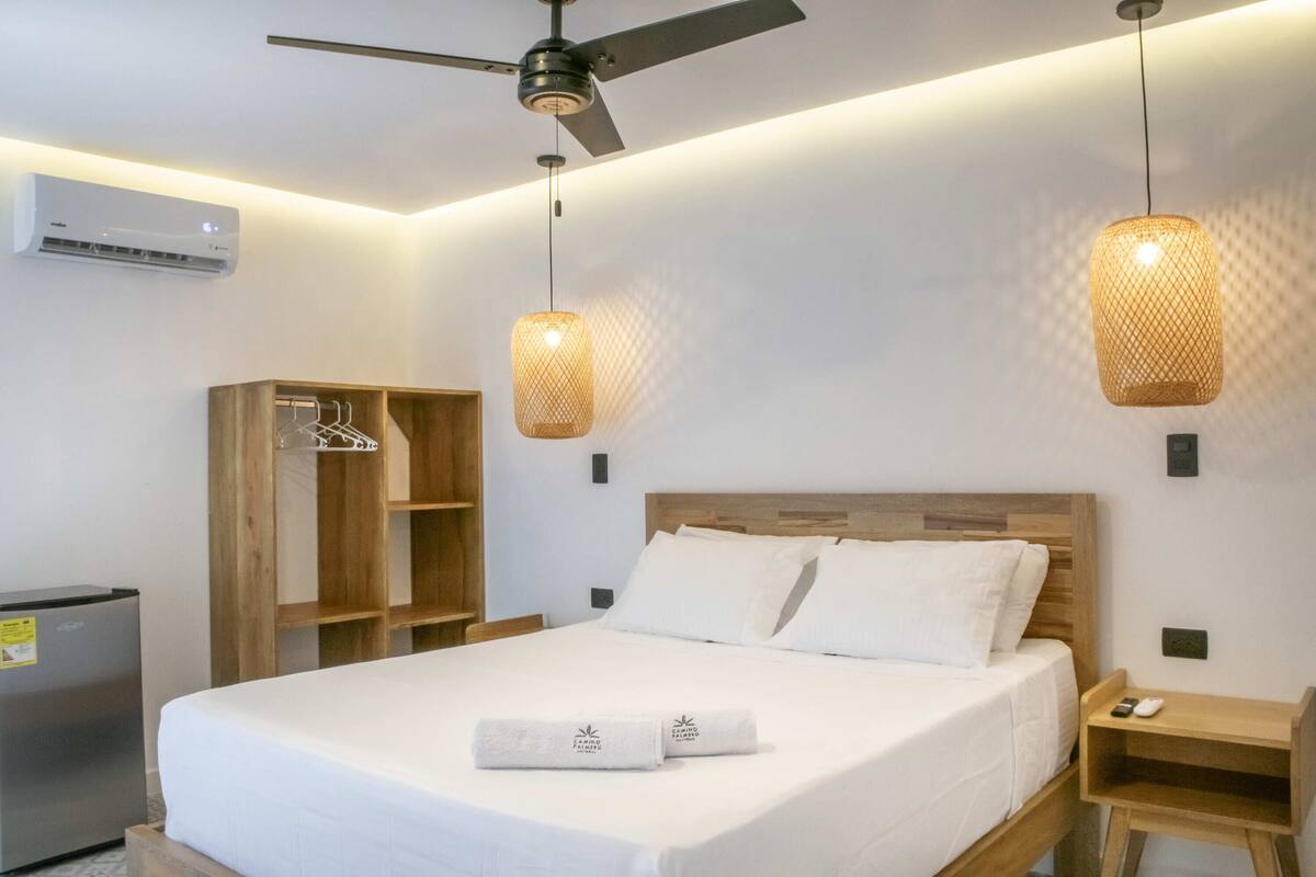 Ein Hotelzimmer mit Bett, Holz-Kopfteil, zwei Pendelleuchten, einem Kleiderschrank, einem Nachttisch und einer Klimaanlage.