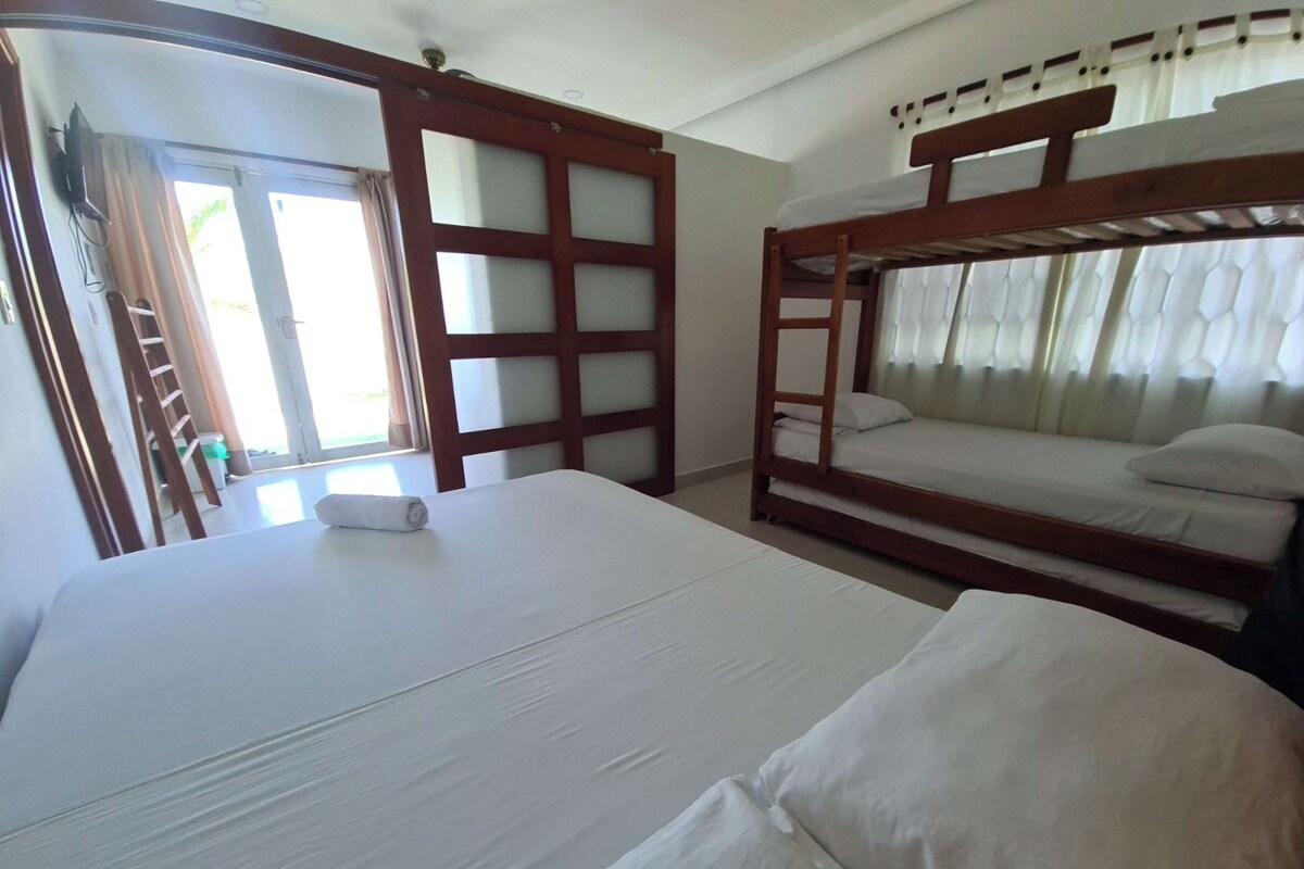 A room with a large bed, a smaller bed, and a bunk bed. There is a TV mounted on the wall and a window with curtains.