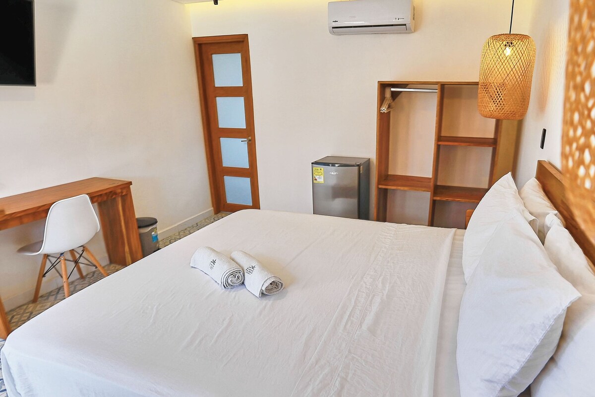 Una cama bien hecha con ropa blanca, un escritorio de madera con una silla, un refrigerador y un armario.