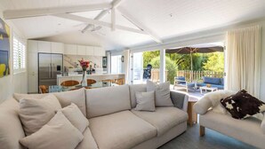 A modern living room with a sofa, a coffee table, and a dining area with chairs. There is a kitchen in the background with white cabinets and a refrigerator. - The Island Veranda - Our Island (Waiheke Island)
