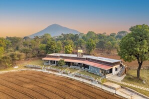 A modern house with a curved roof, surrounded by a fenced property, and a mountain in the background. - Indulge Udaipur By Pack (Udaipur)