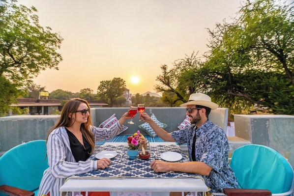 Two people toasting at a table with a patterned tablecloth, surrounded by greenery and a sunset in the background. - Indulge Udaipur By Pack (Udaipur)
