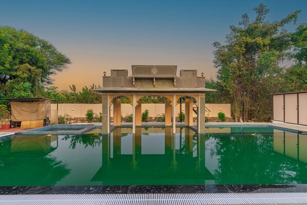 A swimming pool with a central structure, surrounded by greenery and a clear sky. - Indulge Udaipur By Pack (Udaipur)
