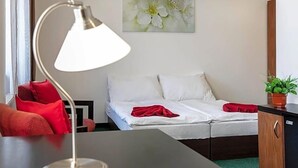 Una habitació d'hotel amb un llit, una taula de treball, una làmpada, una cadira vermella, una planta en test i un quadre a la paret.