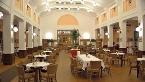 Una àmplia sala de dinar amb diverses taules i cadires, sostres alts i grans finestres.
