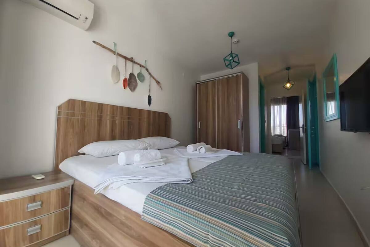 Un dormitorio moderno con cabecera de madera, cama con cubrecama a rayas, mesita de noche, armario y televisor montado en la pared.