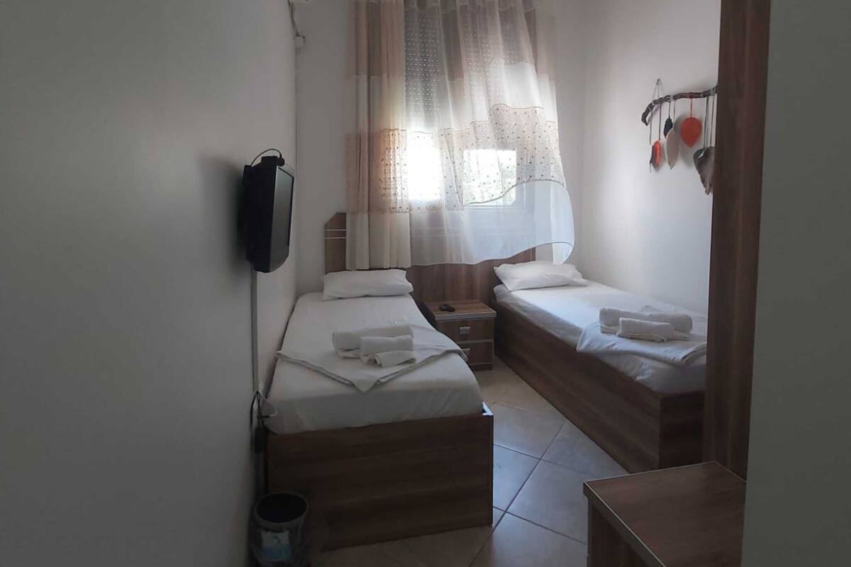 Una habitación pequeña y limpia con dos camas, una mesita de noche, un televisor y una ventana con cortinas.