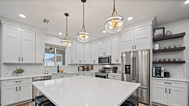 Une cuisine moderne avec des meubles blancs, un grand îlot central, des appareils en acier inoxydable et des luminaires suspendus.