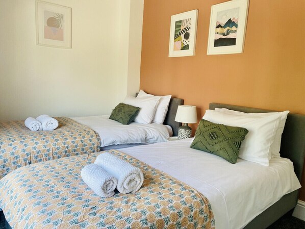 A hotel room with two beds, each with patterned bedding and pillows, a bedside table with a lamp, and artwork on the walls.