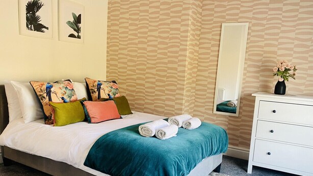 Una cama bien hecha con una colcha color turquesa, varios almohadones y toallas enrolladas.