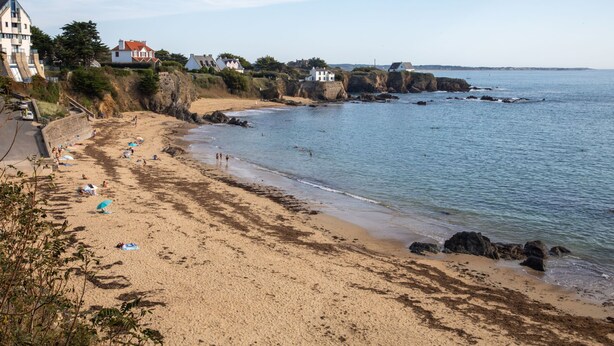 Ein Küstenstrand mit sandigem Ufer, Felsformationen und Gebäuden im Hintergrund.