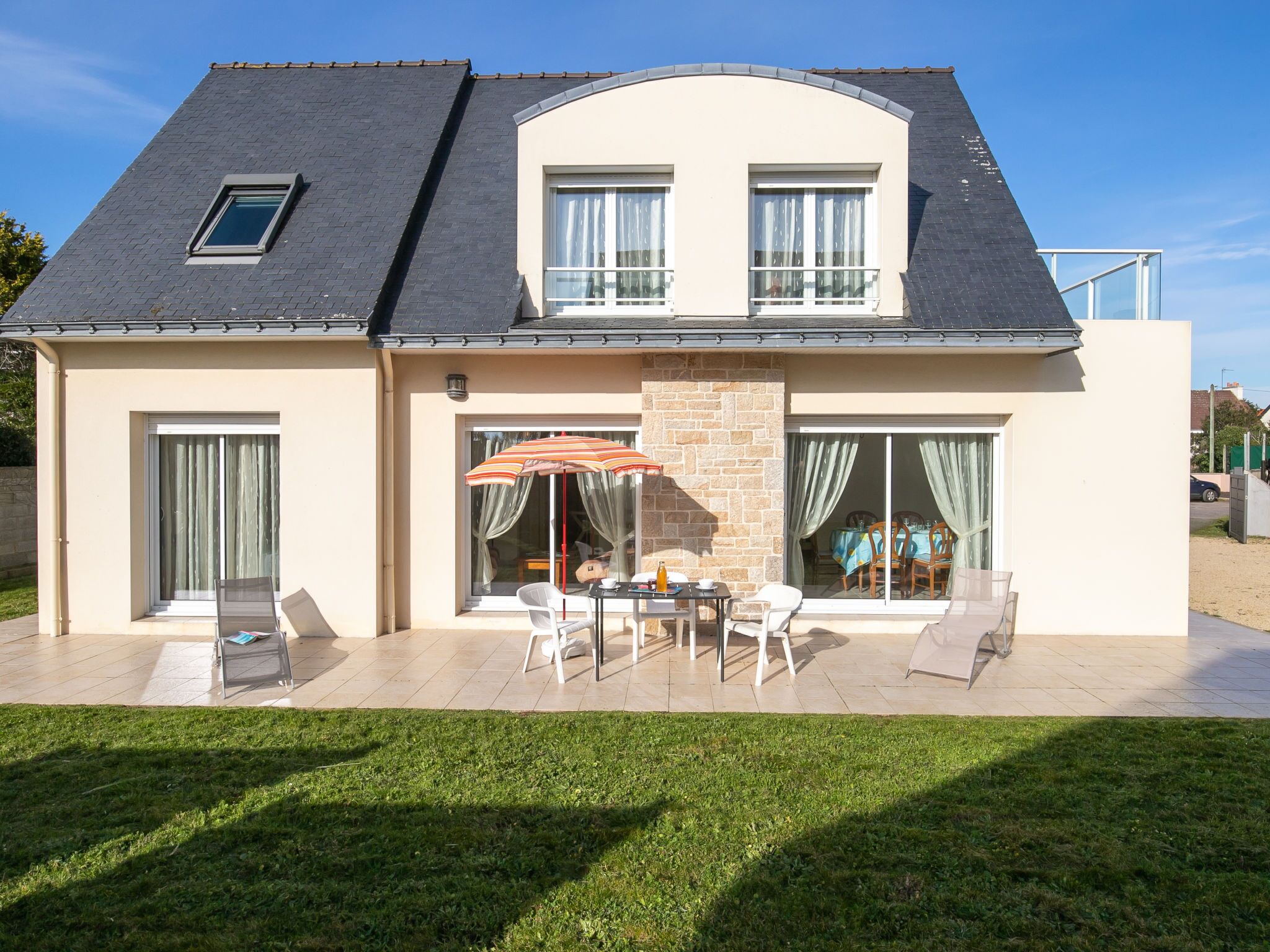 Une maison à deux étages dotée d’un patio comprenant une table et des chaises, un petit jardin et un espace extérieur couvert aménagé en coin salon.