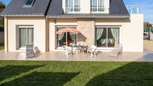 Ein zweistöckiges Haus mit einer Terrassenfläche, die einen Tisch und Stühle, einen kleinen Garten und eine überdachte Sitzgelegenheit bietet.