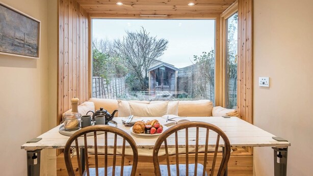 A cozy dining area with a wooden table, two chairs, and a view of a wooden house through large windows.