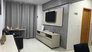 A modern hotel room with a flat-screen TV mounted on the wall, a white console table, a black chair, and a wooden door. - Foz do Iguaçu Belém Residential Apartment located upper floor 10' FROM COP30 (Pedreira Belém)