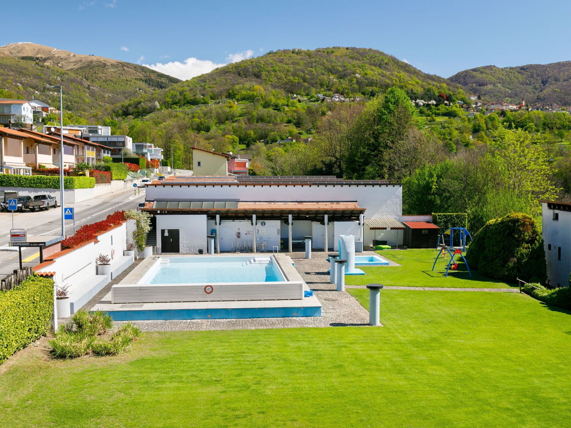 A modern building with a swimming pool, surrounded by a well-maintained lawn and a view of a hilly landscape.