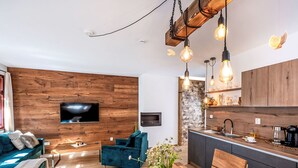 A modern living room with a wooden wall, a TV mounted above a sofa, a dining area with a table and chairs, and a kitchen with a stone backsplash and wooden cabinets. - Apartment Lipno by Interhome (Horni Plana)