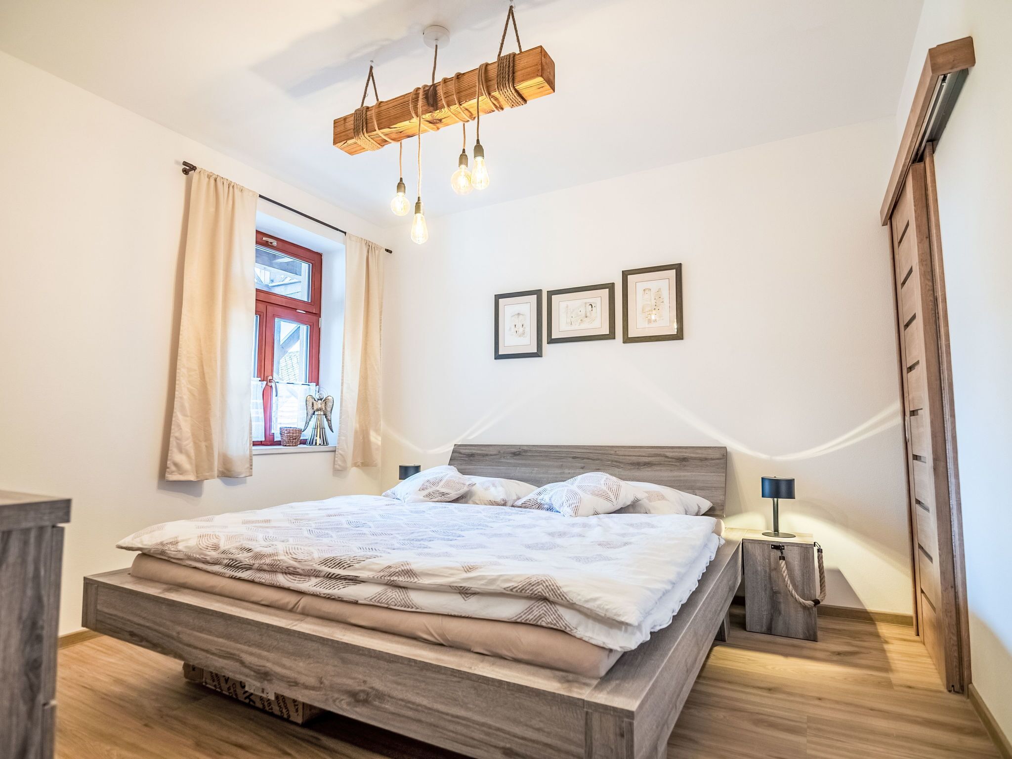 A bedroom with a wooden bed, a window with curtains, a bedside table with a lamp, and framed pictures on the wall.