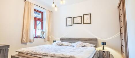 A bedroom with a wooden bed, a window with curtains, a bedside table with a lamp, and framed pictures on the wall.