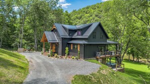 Een groot, donker huis met een brede veranda en terras, omringd door weelderige begroeiing en bomen.