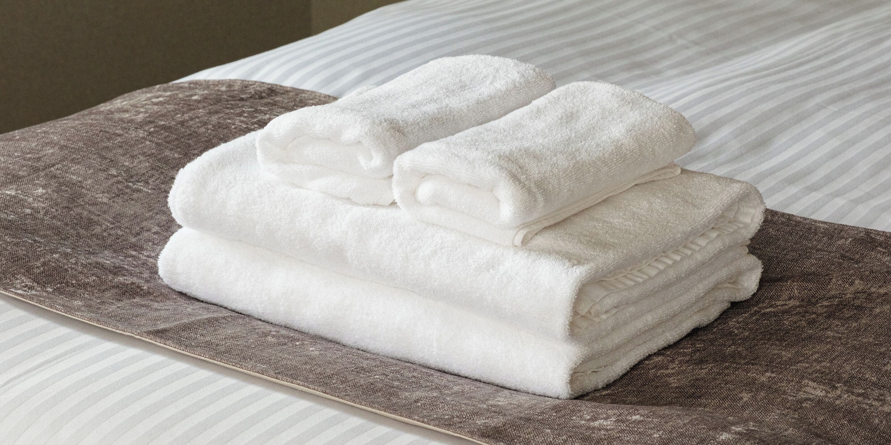 Una pila de toallas blancas ordenadas sobre una bandeja marrón con textura, sobre una cama con ropa de cama a rayas.