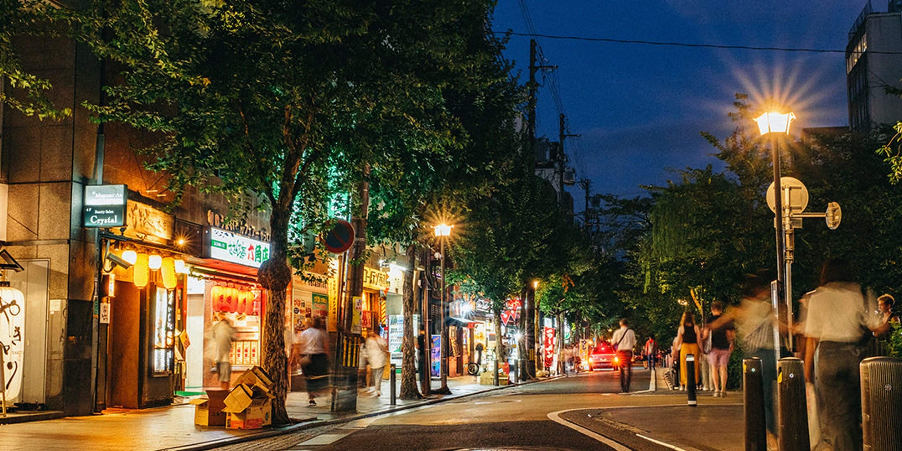 Una calle iluminada de noche con escaparates brillantes y gente caminando.