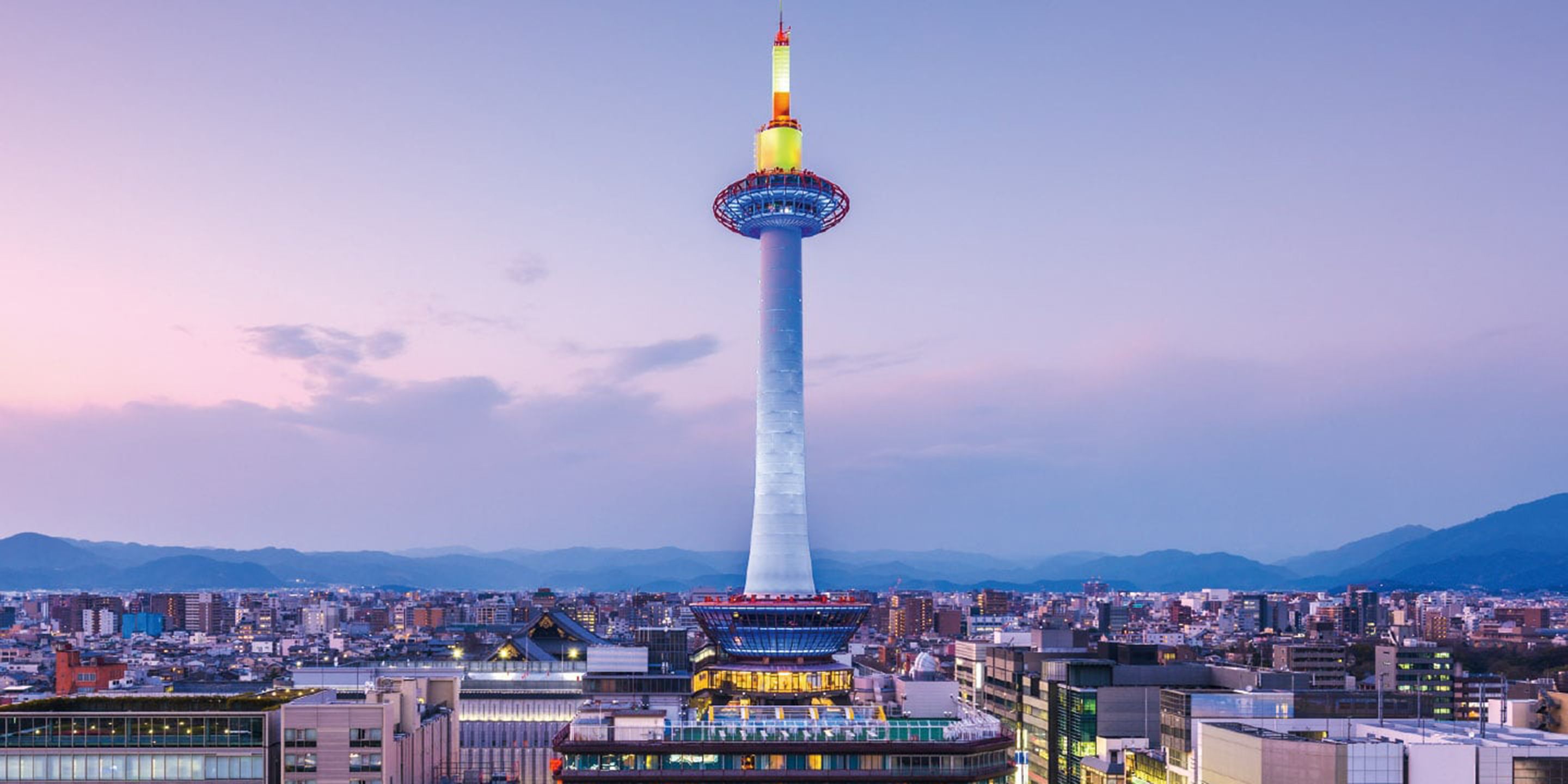A tall tower with a spire, surrounded by a cityscape and mountains in the background.