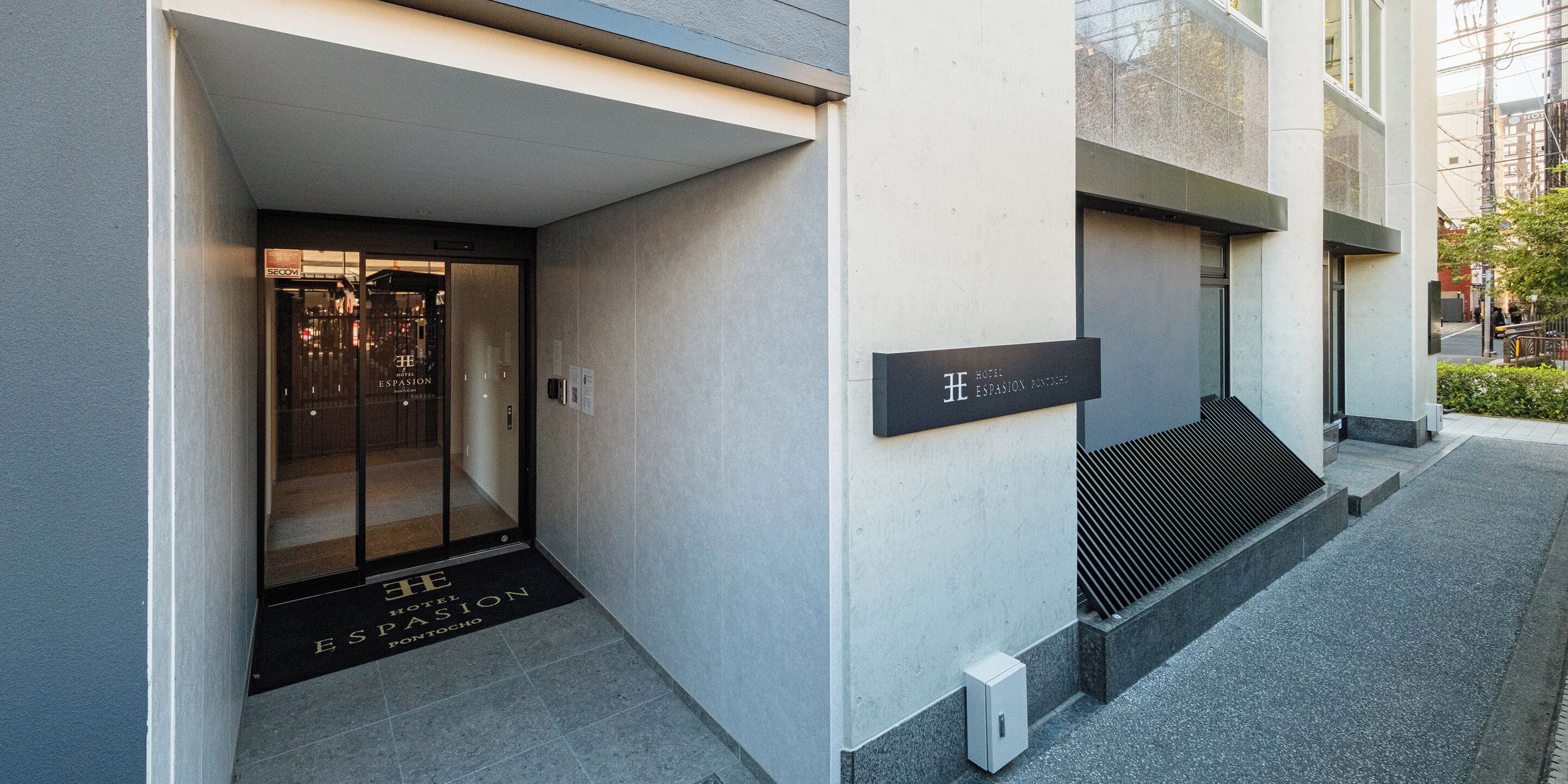 Der Eingang des Hotels ESPASION PONTOCHO mit modernem Design, darunter eine Glastür und eine schwarze Schilder.'