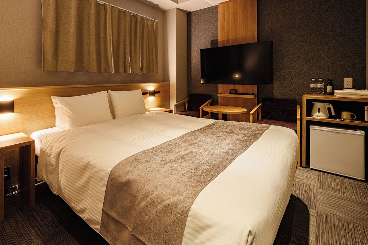 Habitación de hotel con cama, mesitas de noche, televisor, una mesita pequeña y un mini refrigerador.